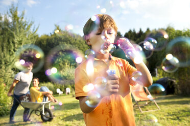 Happy family in garden with man pushing woman in wheelbarrow and boy blowing soap bubbles - TOYF001481