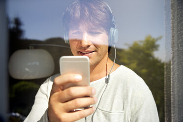 Man behind windowpane listening to music from smartphone - TOYF001485