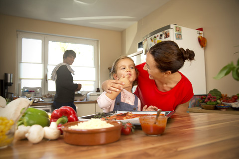 Glückliche Mutter und Tochter in der Küche bei der Zubereitung einer Pizza mit dem Vater im Hintergrund, lizenzfreies Stockfoto