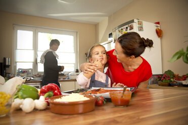 Happy mother and daughter in kitchen preparing pizza with father in background - TOYF001514