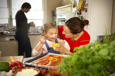 Happy mother and daughter in kitchen preparing pizza with father in background - TOYF001515