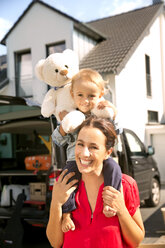 Mutter, die ihren Sohn mit einem Teddybär auf den Schultern trägt, steht vor dem Haus - TOYF001522