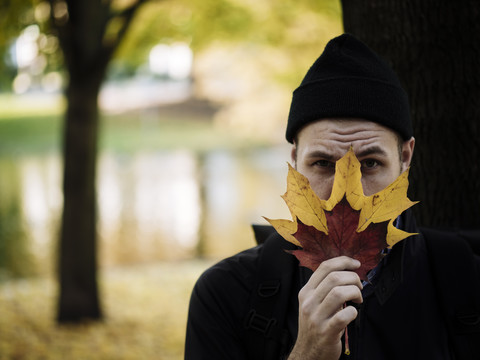 Porträt eines Mannes mit Wollmütze, der sich hinter Herbstblättern versteckt, lizenzfreies Stockfoto