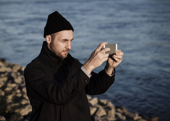 Germany, Cologne, man wearing autumn fashion standing in front of Rhine River taking a selfie - DASF000023