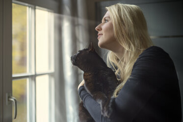 Woman and her cat looking through the window at home - FRF000354