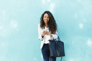 Portrait of smiling young businesswoman with smartphone and leather bag in front of a blue wall - EBSF001018