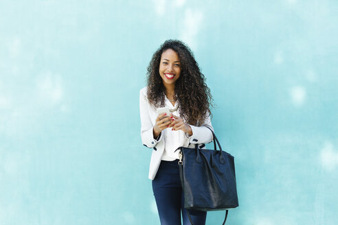 Porträt einer lächelnden jungen Geschäftsfrau mit Smartphone und Ledertasche vor einer blauen Wand - EBSF001018
