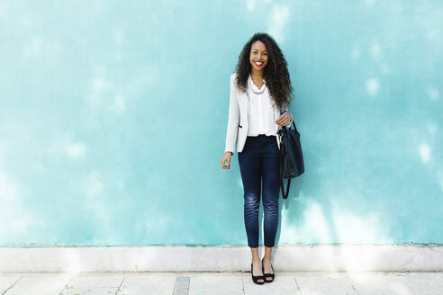 Porträt einer lächelnden jungen Geschäftsfrau mit Ledertasche, die vor einer blauen Wand steht - EBSF001019