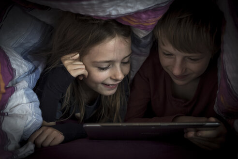 Bruder und Schwester liegen Seite an Seite und benutzen ein digitales Tablet unter einer Decke - SARF002301