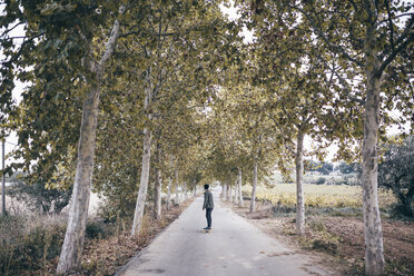 Spain, Tarragona, back view of young man with longboard on autumnal country road - JRFF000176