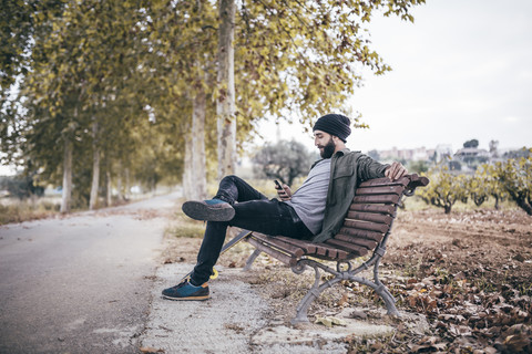 Spanien, Tarragona, junger Mann sitzt auf einer Bank an einer herbstlichen Landstraße und schaut auf sein Smartphone, lizenzfreies Stockfoto