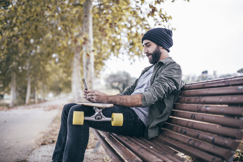 Spanien, Tarragona, junger Mann sitzt auf einer Bank mit seinem Longboard und macht ein Selfie mit Smartphone - JRFF000189