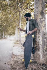 Spanien, Tarragona, junger Mann mit Longboard lehnt an einem Baumstamm und schaut auf sein Smartphone - JRFF000190