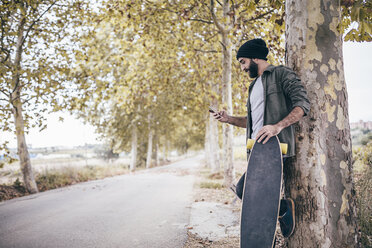 Spanien, Tarragona, junger Mann mit Longboard lehnt an einem Baumstamm und schaut auf sein Smartphone - JRFF000191