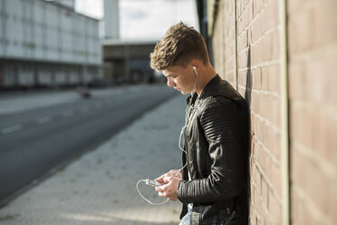 Junger Mann mit Ohrstöpseln, der auf sein Mobiltelefon schaut - UUF005933