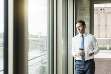 Portrait of businessman with coffee to go and smartphone looking through window - UUF005968