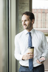 Portrait of smiling businessman with coffee and smartphone looking through window - UUF005969