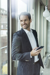 Portrait of smiling businessman with smartphone looking through window - UUF005976