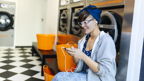 Lächelnde junge Frau hört Musik mit Kopfhörern in einem Waschsalon, lizenzfreies Stockfoto