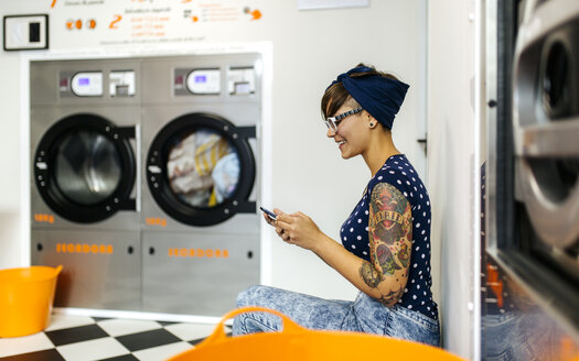 Tätowierte junge Frau schaut in einem Waschsalon auf ihr Smartphone - MGOF001035