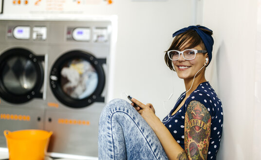 Porträt einer tätowierten jungen Frau, die mit Kopfhörern in einem Waschsalon Musik hört - MGOF001036