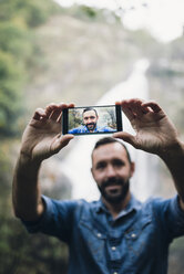Bärtiger Mann macht ein Selfie mit Smartphone vor einem Wasserfall - RAEF000650