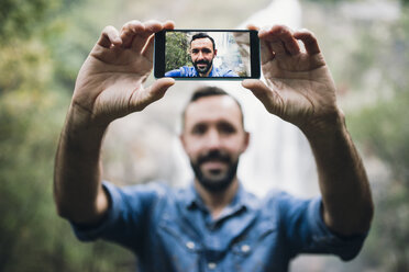 Bearded man taking a selfie with smartphone in front of a waterfall - RAEF000651