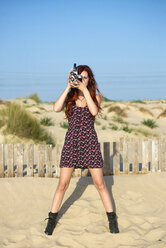 Spain, El Puerto de Santa Maria, young woman using old film camera on the beach - KIJF000027