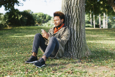 Young man with smartphone leaning against tree trunk on a meadow - EBSF001055