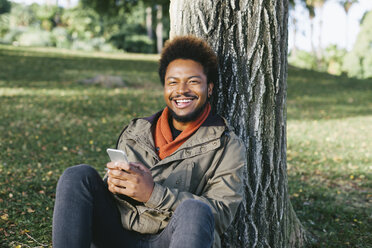 Porträt eines lächelnden jungen Mannes mit Smartphone, der sich gegen einen Baumstamm lehnt - EBSF001056