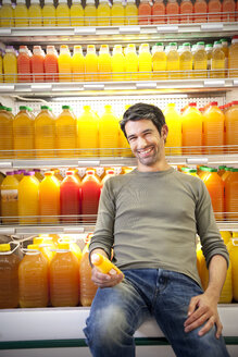 Portrait of smiling man sitting in front of fridge with rows of juice bottles in a supermarket - RMAF000223