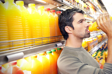 Man drinking a bottle of orange juice in front of fridge in a supermarket - RMAF000225