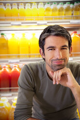 Porträt eines lächelnden Mannes, der in einem Supermarkt vor einem Kühlschrank mit Reihen von Saftflaschen sitzt - RMAF000226