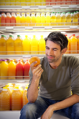 Porträt eines lächelnden Mannes, der in einem Supermarkt vor einem Kühlschrank mit Reihen von Saftflaschen sitzt und eine Orange hält, lizenzfreies Stockfoto