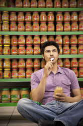 Portrait of man sitting on the floor of a supermarket tasting glass of peanut butter - RMAF000240