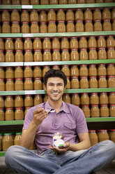 Smiling man sitting on the floor of a supermarket tasting glass of chocolate spread - RMAF000244