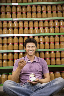 Lächelnder Mann, der in einem Supermarkt auf dem Boden sitzt und ein Glas Schokoladenaufstrich probiert - RMAF000244