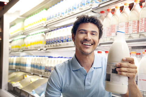 Porträt eines lächelnden Mannes, der in einem Supermarkt vor einem Kühlschrank sitzt und eine Milchflasche hält, lizenzfreies Stockfoto