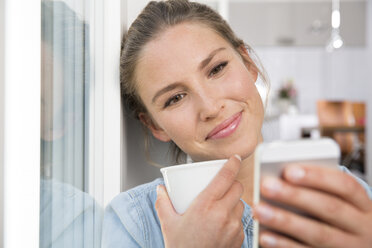 Young woman sitting at the window, drinking coffee an reading sms - FKF001550