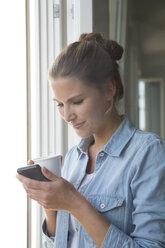 Junge Frau steht mit Smartphone und Kaffeetasse am Fenster - FKF001554