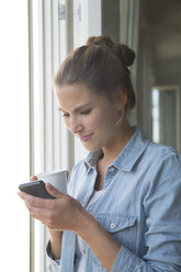 Young woman standing at the window with smart phone and coffee cup - FKF001554