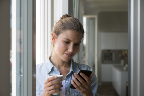 Junge Frau steht mit Smartphone und Kaffeetasse am Fenster, lizenzfreies Stockfoto