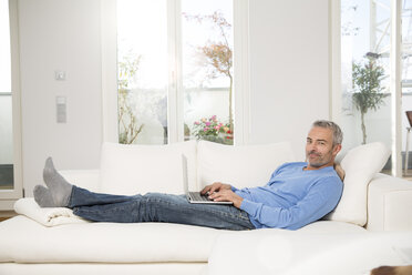 Mature man at home, sitting on couch, using laptop - FKF001577