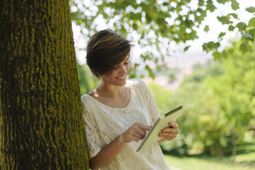 Lächelndes Teenager-Mädchen lehnt an einem Baumstamm und benutzt ein digitales Tablet - GIOF000517