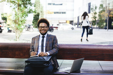 Lächelnder junger Geschäftsmann mit Laptop und Notizbuch auf einer Bank - EBSF001100