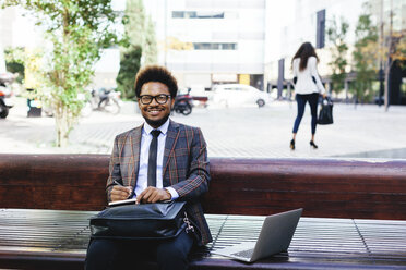 Smiling young businessman with laptop and notebook on bench - EBSF001100