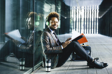 Smiling young businessman with headphones and folder sitting outdoors - EBSF001123