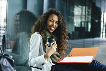 Smiling young businesswoman with coffee to go mug and folder sitting outdoors - EBSF001129