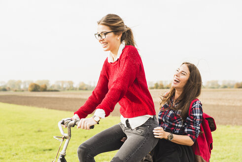 Zwei glückliche junge Frauen teilen sich ein Fahrrad in ländlicher Umgebung - UUF006040