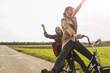 Two exuberant young women sharing a bicycle in rural landscape - UUF006044